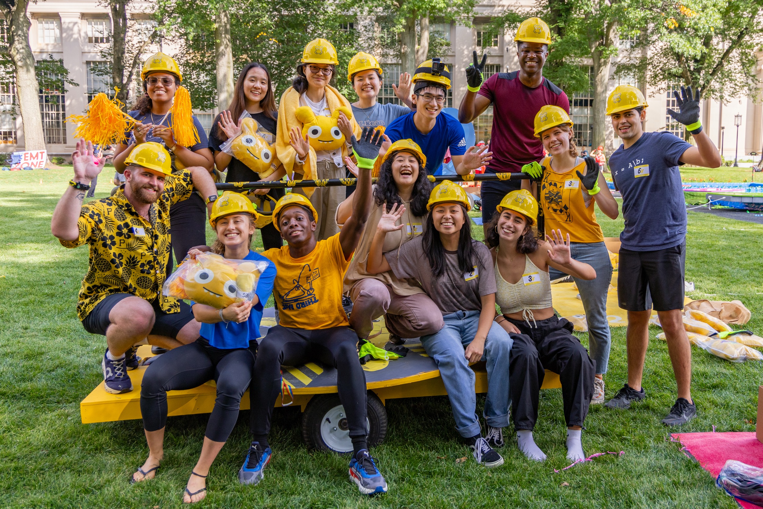 A group of smiling people wearing yellow construction hats and yellow or blue clothing pose together outdoors on grass, waving at the camera. Some hold plush toys, and colorful decorations are visible around them.