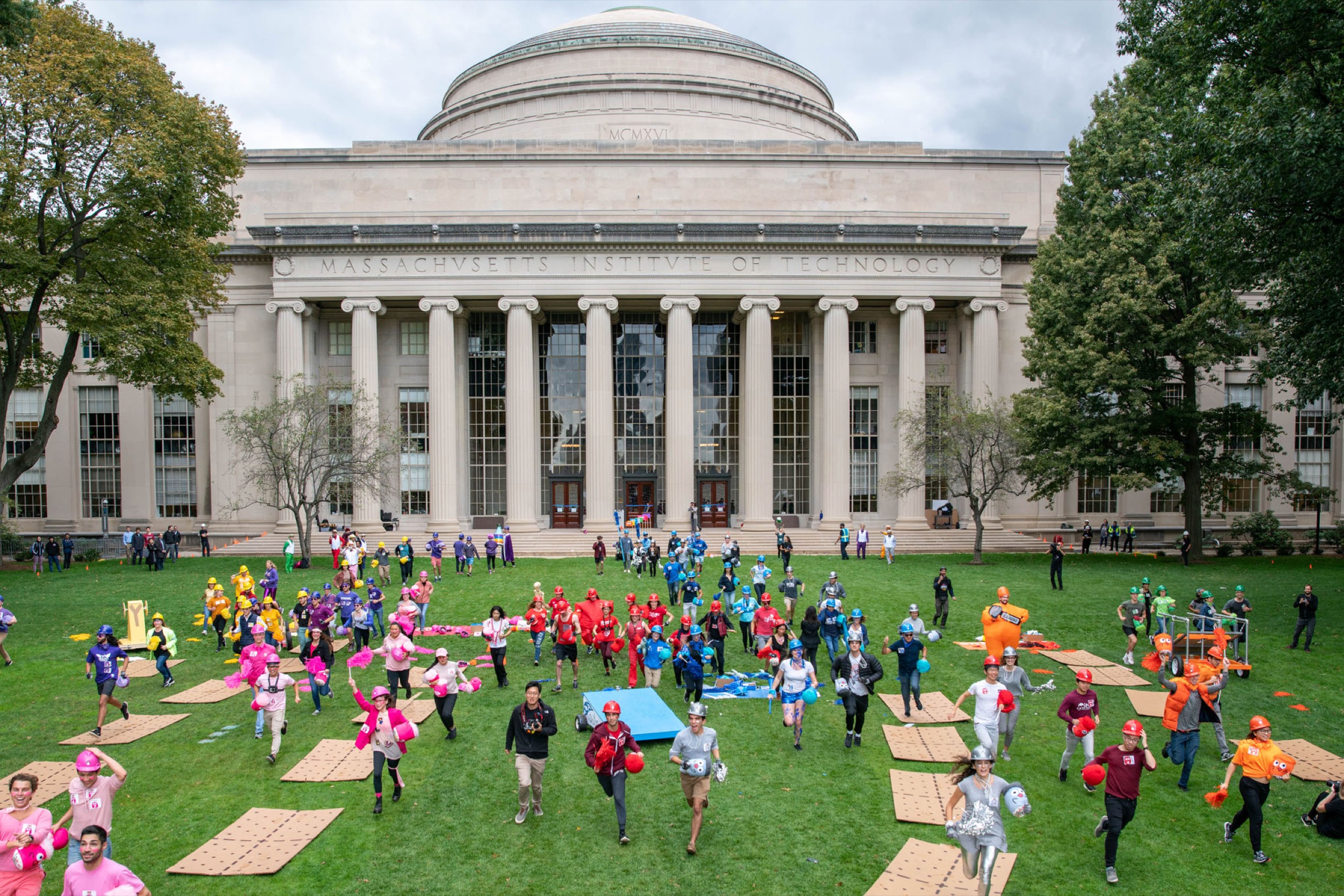 A large group of people in colorful shirts run across the grass in front of the domed Maclaurin building at the Massachusetts Institute of Technology, with cardboard pieces scattered on the lawn.