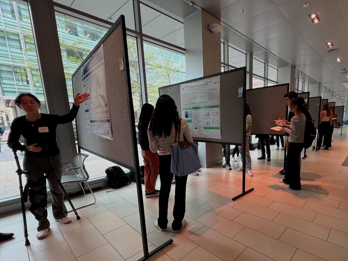 Several people stand in a bright hallway, presenting and discussing research posters displayed on large boards. Some participants point to their posters while others listen and take notes.