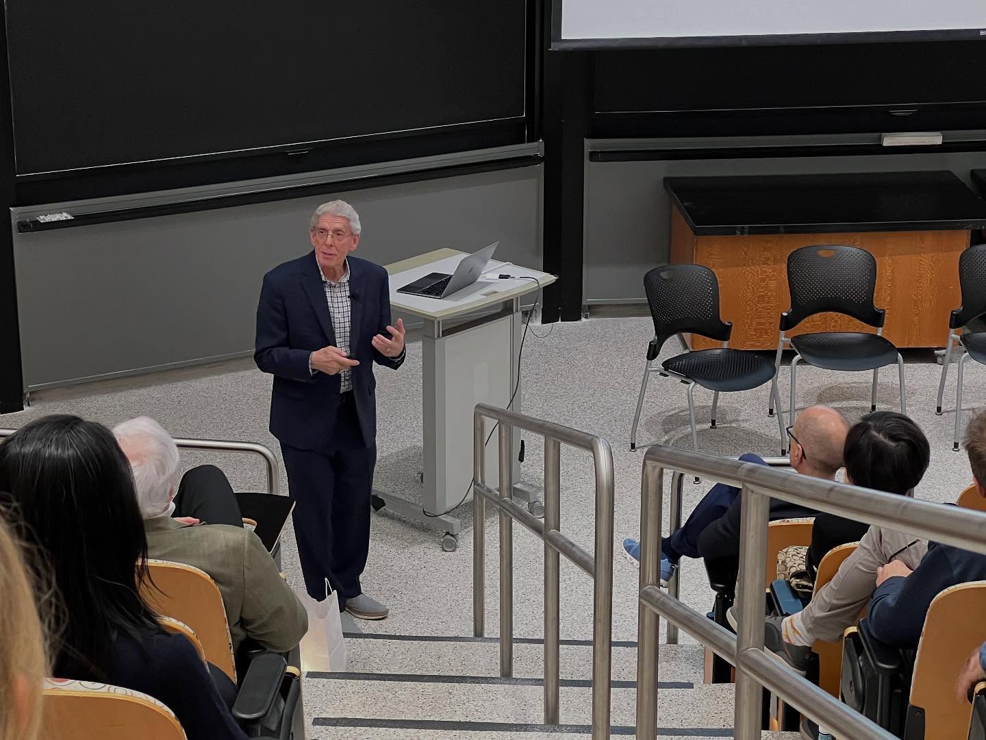 A man in a suit stands at a podium giving a lecture to an audience in a tiered classroom. Several people sit in rows, listening, with empty chairs and a laptop visible near the speaker.