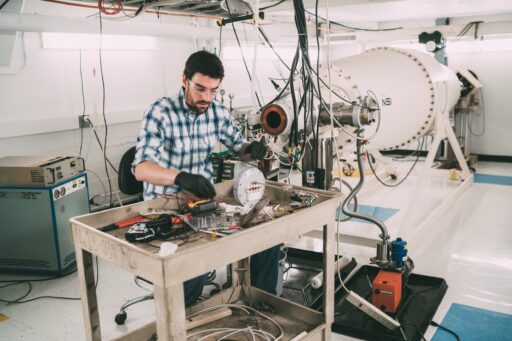 A man wearing glasses and gloves works with electronic equipment on a metal cart in a laboratory, with a large cylindrical scientific instrument and cables in the background.