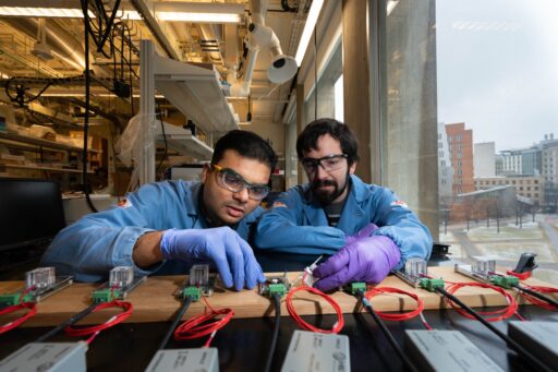 Two scientists wearing blue lab coats, safety glasses, and purple gloves work on electronic equipment with wires at a lab bench near a large window overlooking a campus with buildings outside.