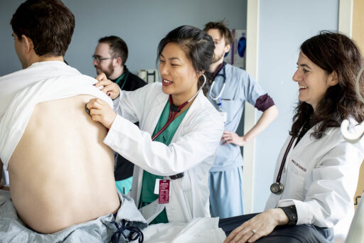 A medical professional examines a patient’s back with a stethoscope while others, also in white coats and scrubs, observe and smile in a clinical setting.