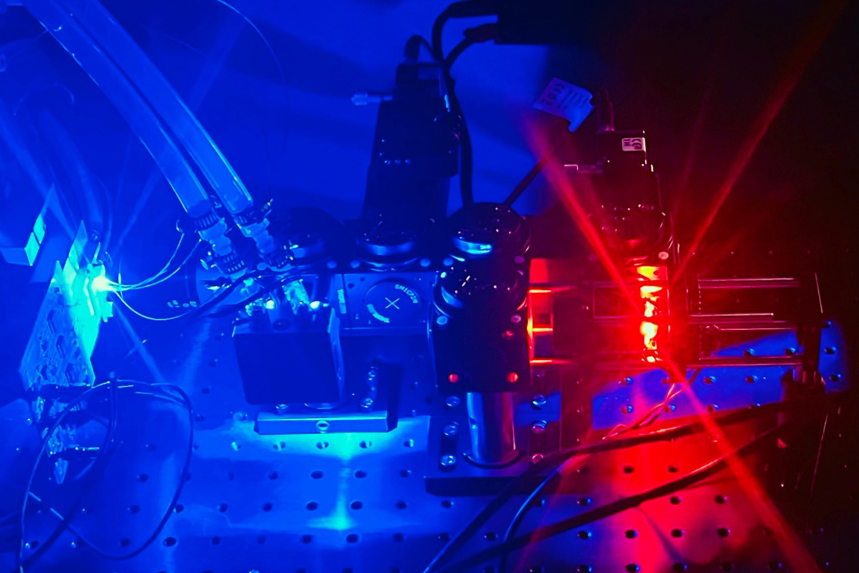Close-up of a scientific optical setup on a metal breadboard, illuminated by blue and red LED lights, showing lenses, mirrors, cables, and electronic components arranged for an experiment.