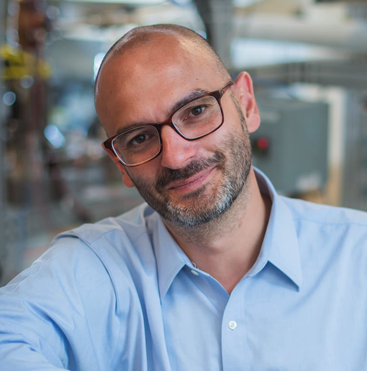 A man with glasses, a beard, and a shaved head, wearing a light blue collared shirt, smiles while leaning slightly forward in an indoor setting with blurred industrial equipment in the background.