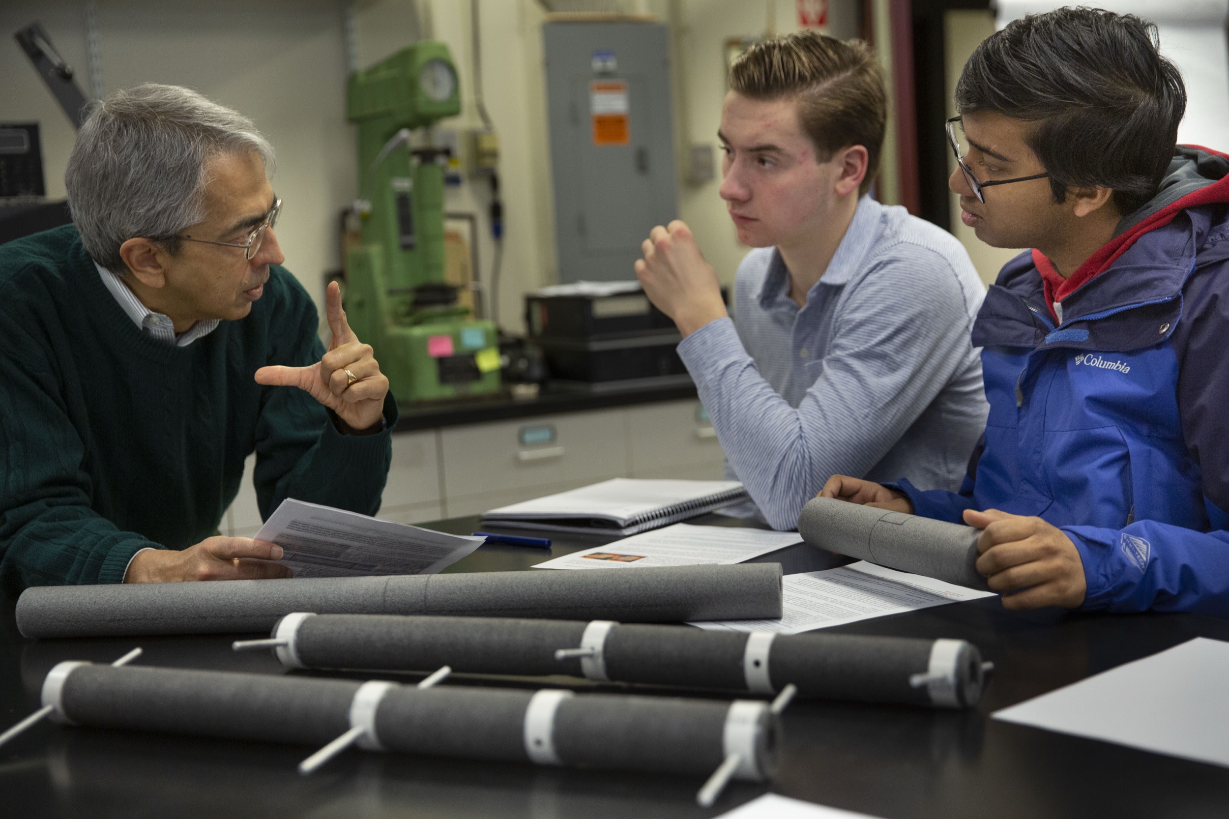 A professor gestures while talking to two students at a lab table. The students listen attentively, holding rolled-up materials. Several cylindrical lab samples are arranged on the table in front of them.