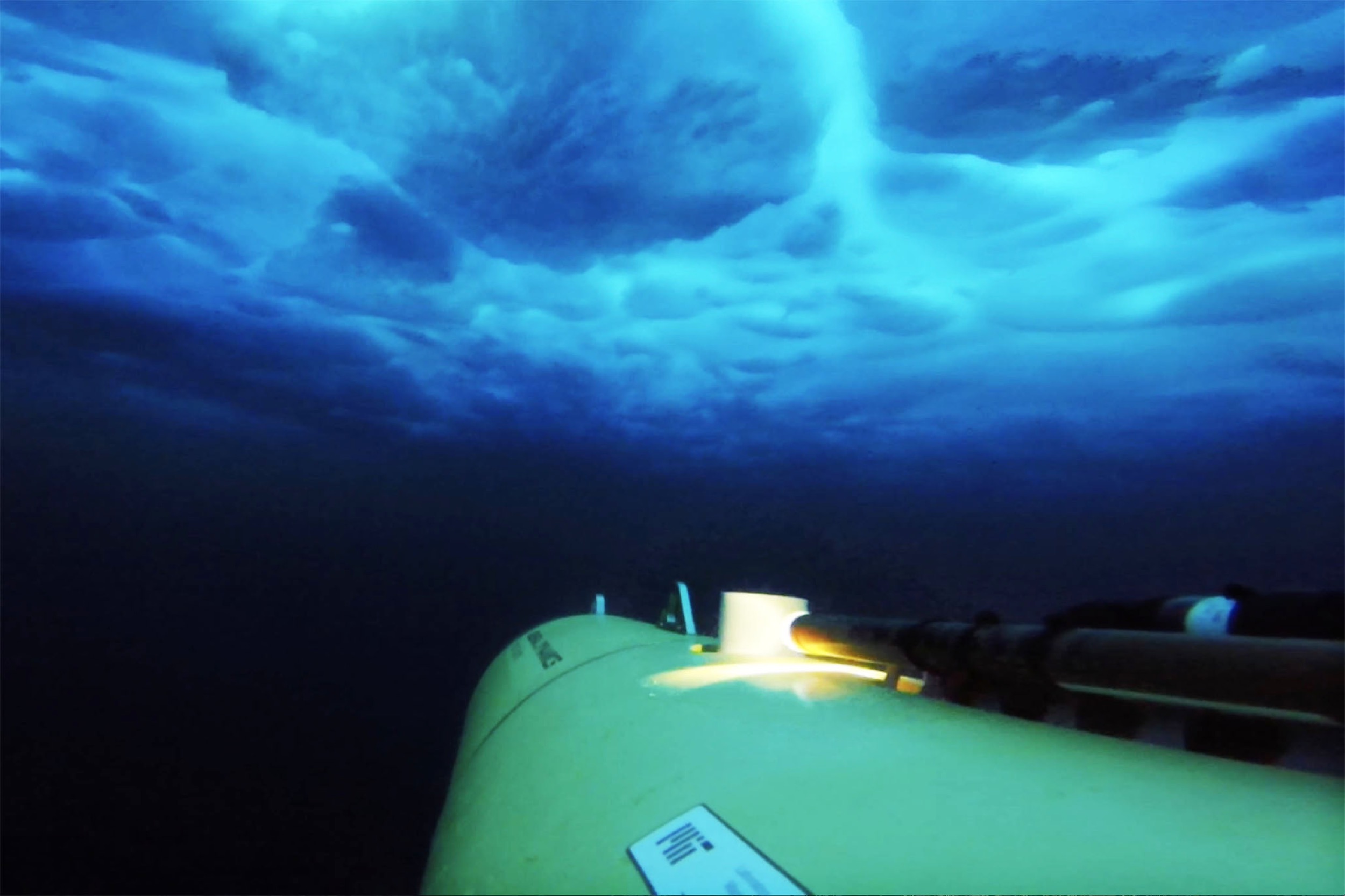 A yellow underwater vehicle navigates deep, dark blue water beneath a layer of ice, illuminated by a blue glow from the ice above and a small onboard light.