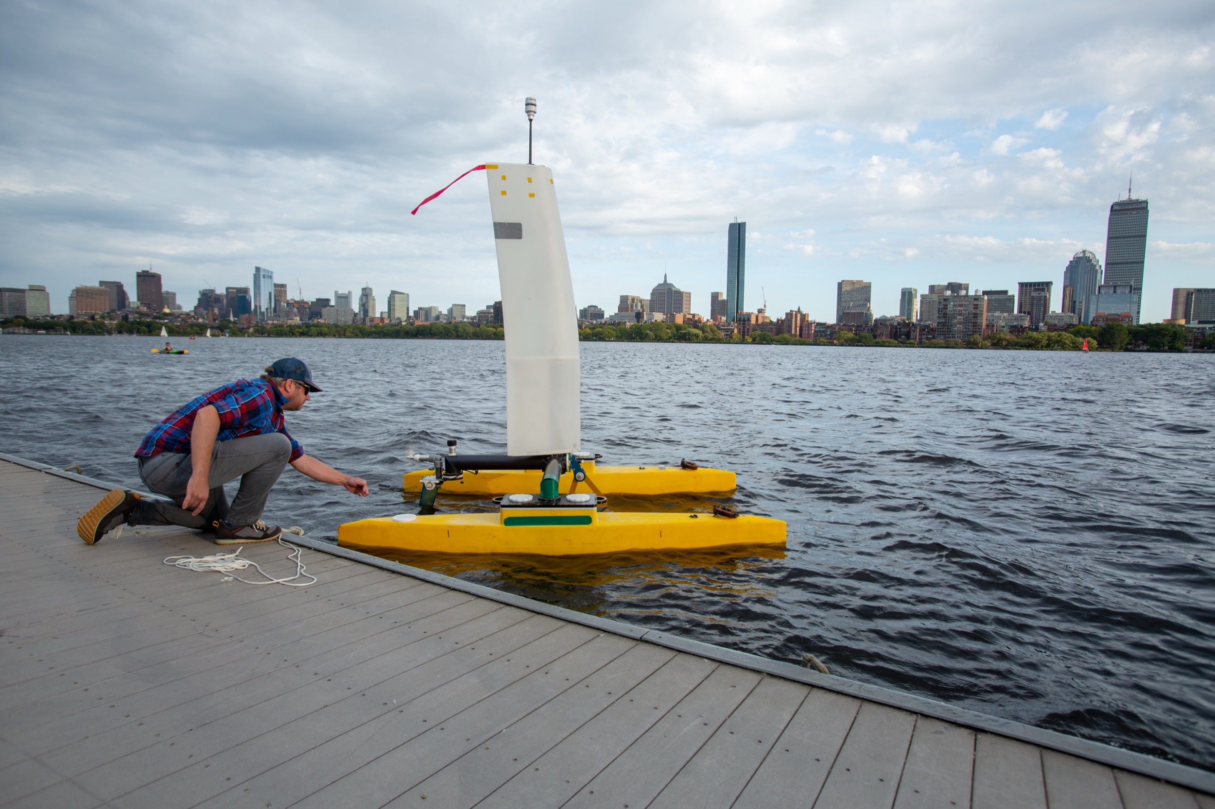 A person kneels on a dock, reaching toward a small, yellow, unmanned sailboat on the water. A city skyline with tall buildings is visible in the background under a partly cloudy sky.