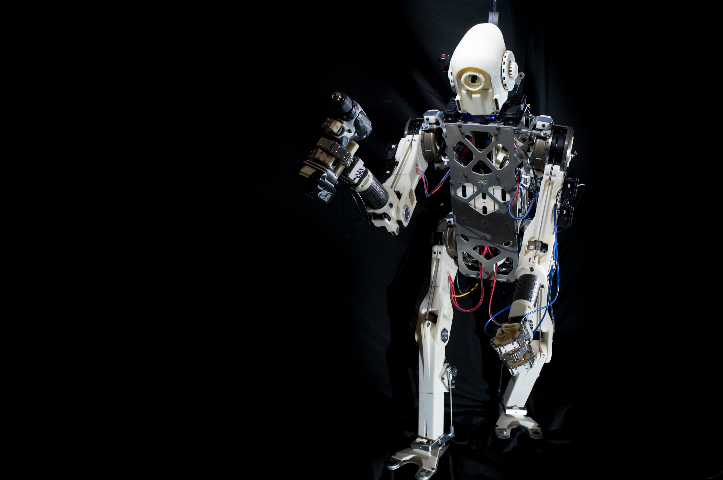A humanoid robot with a white head and metal frame stands against a black background, holding a tool in its right hand. Wires and mechanical components are visible throughout its body.