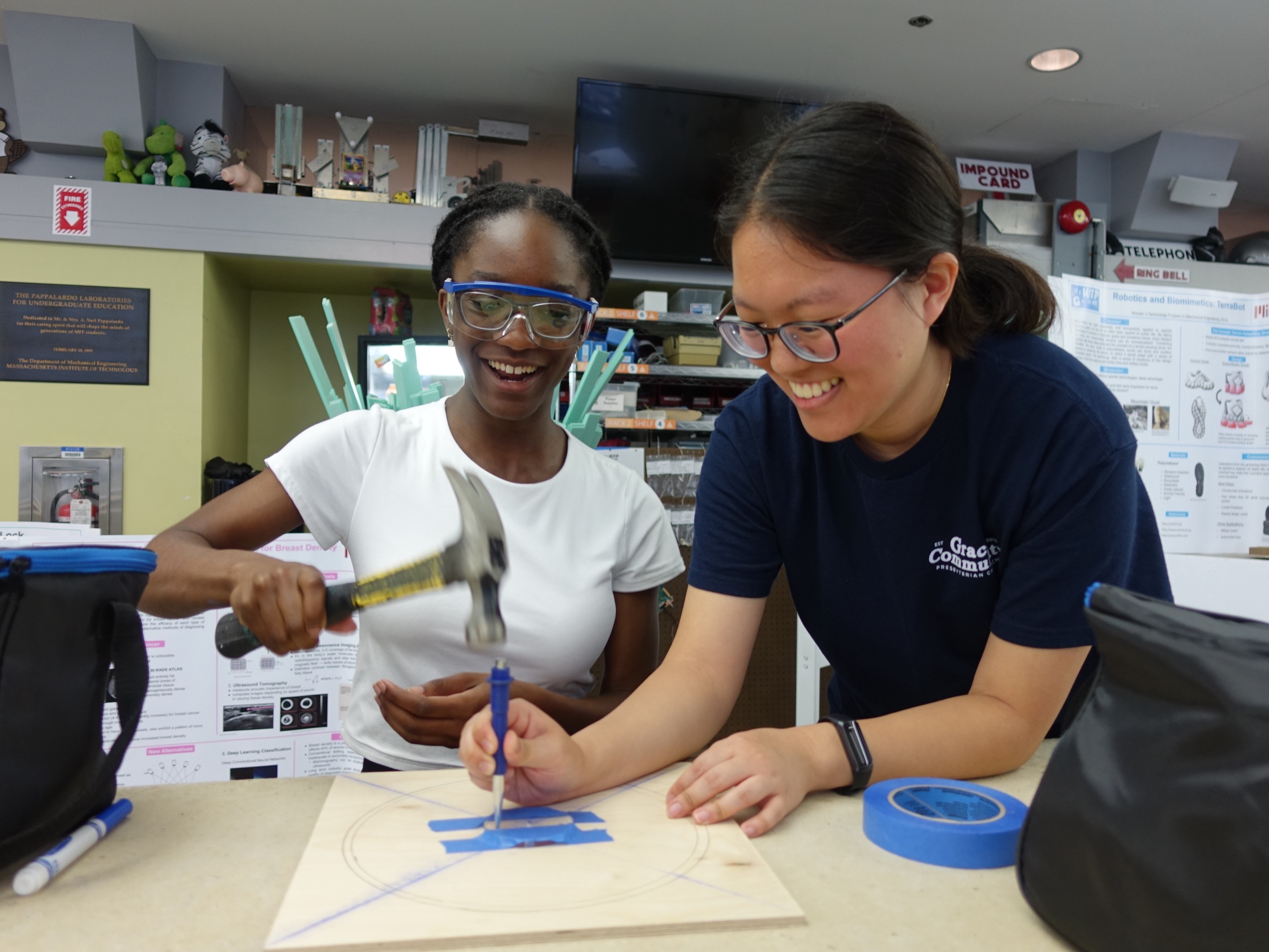 Two women smiling and working together on a project; one is holding a hammer while the other holds a chisel on a piece of wood marked with a circle, surrounded by blue tape and craft supplies on the table.