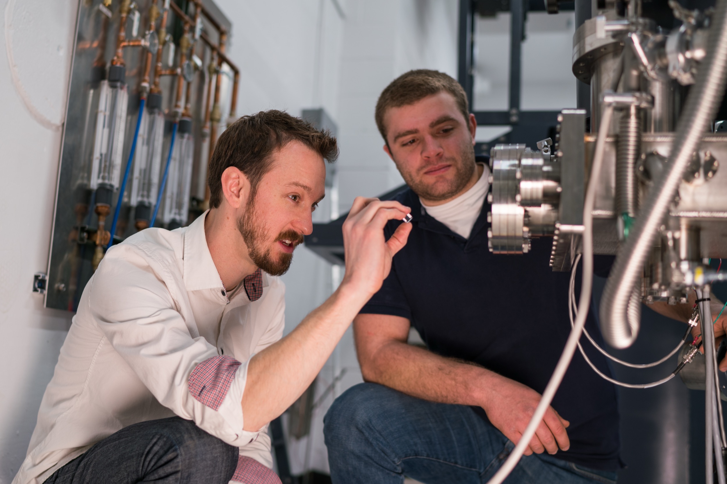 Two men crouch next to complex laboratory equipment. One points towards the machine while explaining something to the other. Both appear focused and engaged in a science or engineering setting.