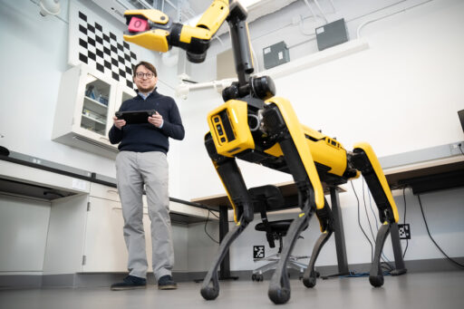 A person stands in a lab holding a tablet, observing a yellow, dog-like robot with four legs and a camera on its head. The setting appears modern with desks, shelves, and technical equipment visible.