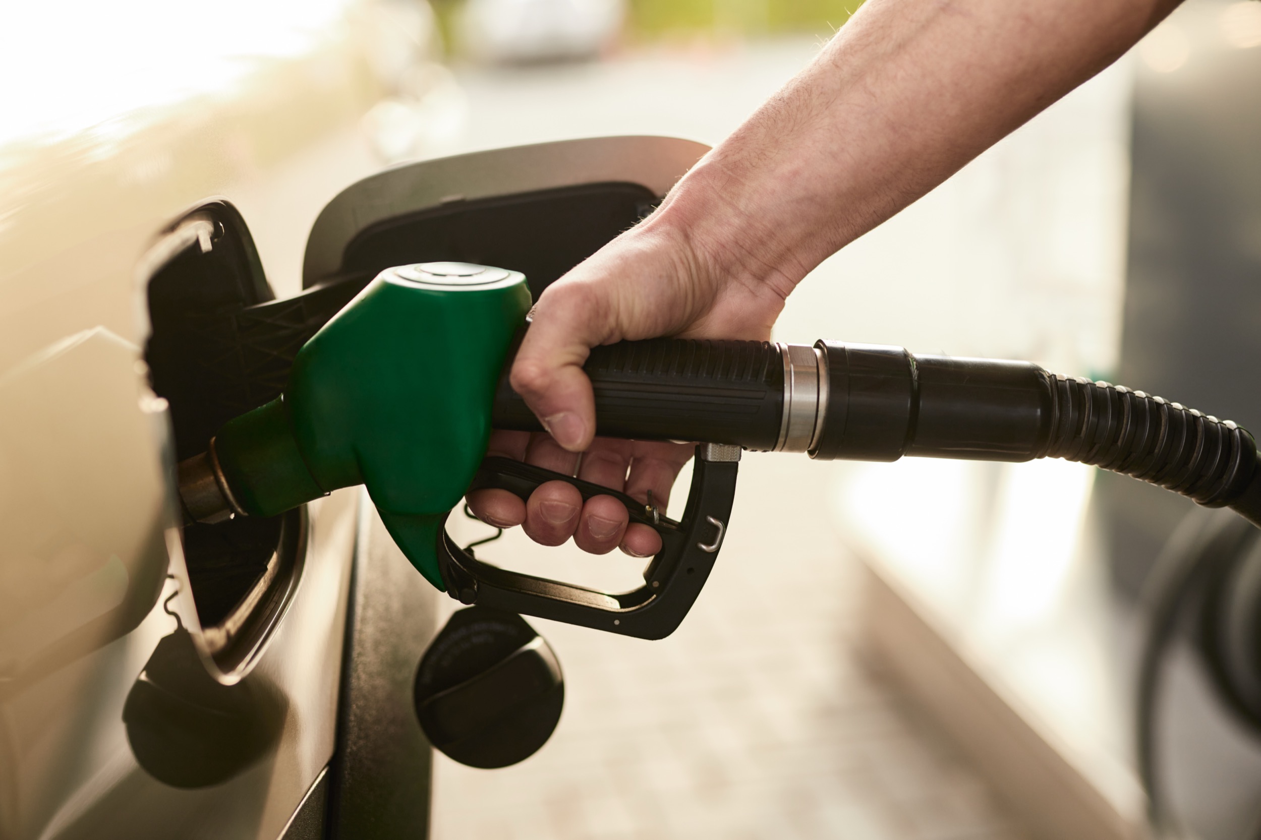 A close-up of a person’s hand holding a green fuel nozzle, refueling a car at a gas station. The cars fuel door is open and the nozzle is inserted into the tank.