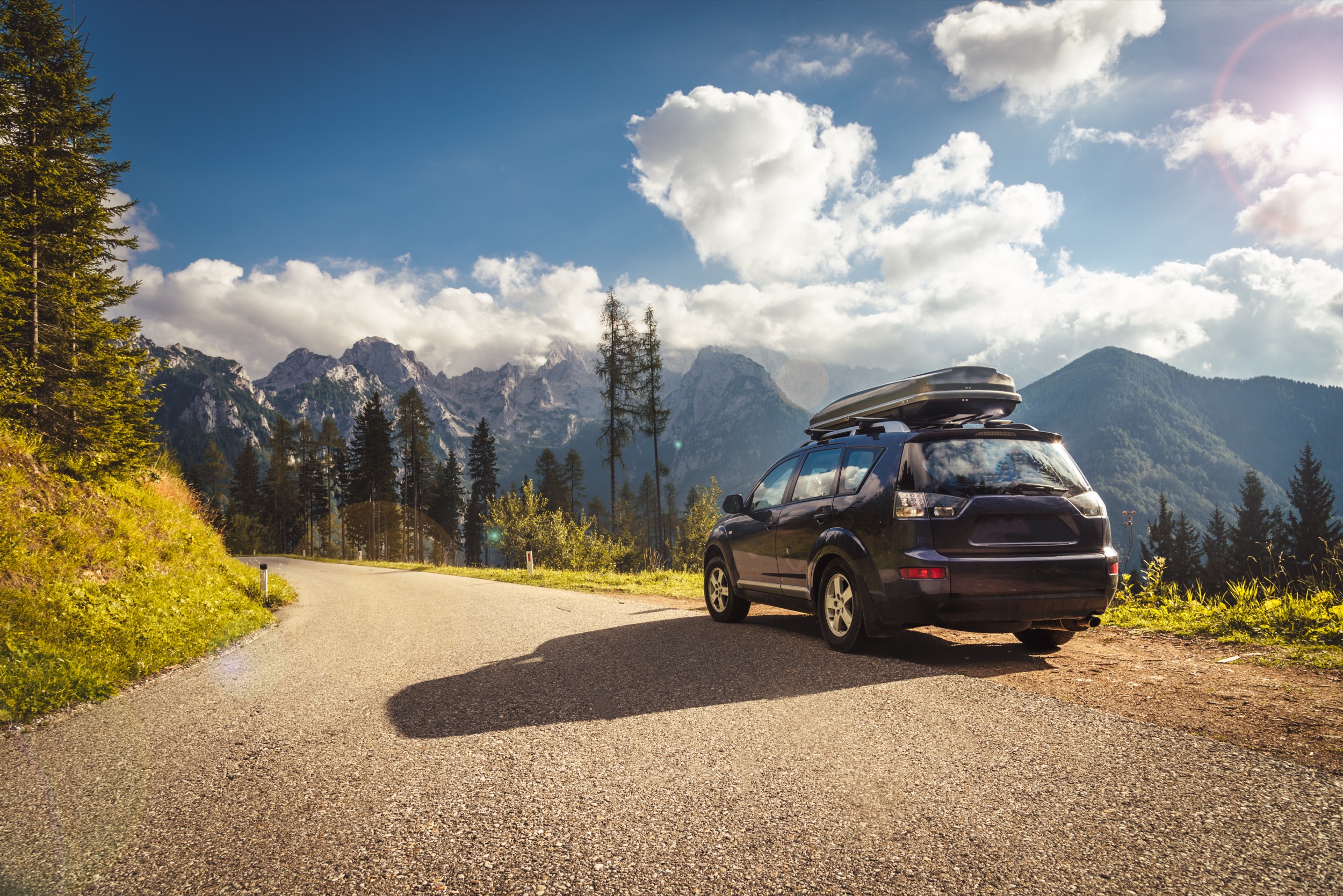 A black SUV with a roof box is parked on a scenic mountain road, surrounded by tall trees, with snow-capped peaks and a bright blue sky with clouds in the background.