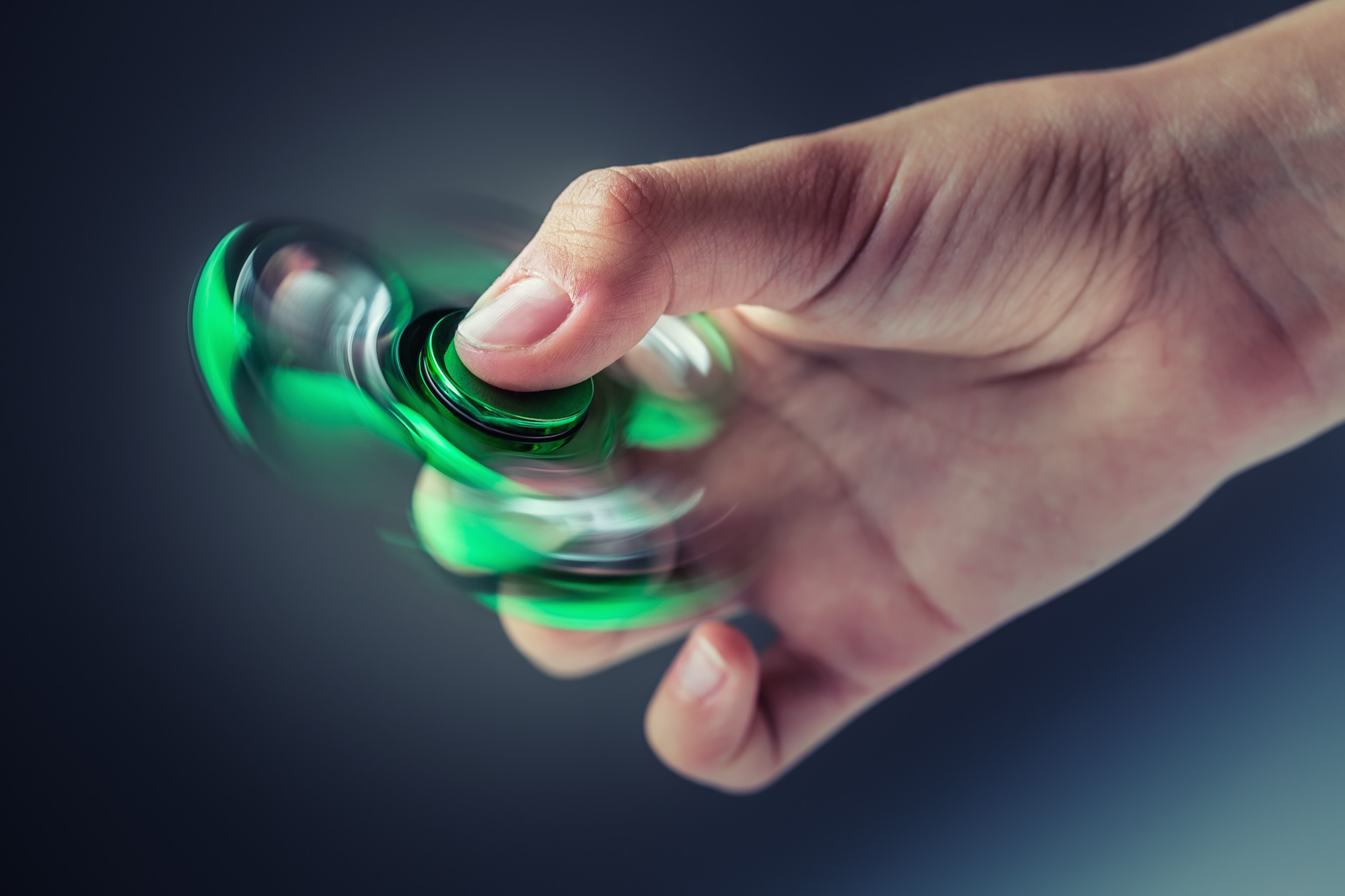 A hand is holding and spinning a green and black fidget spinner, with the motion creating a blurred effect on the spinner’s arms against a dark, blurred background.