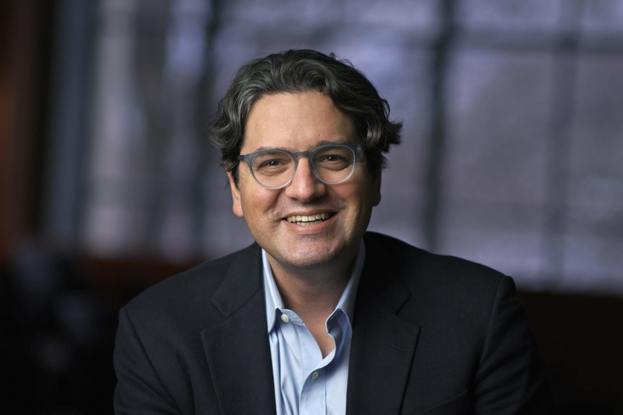 A man with wavy dark hair and glasses smiles while wearing a dark blazer and light blue shirt, seated indoors with a blurred window in the background.