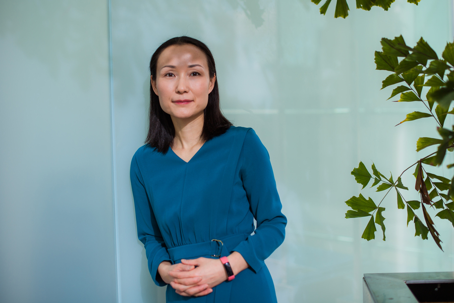 A woman with straight black hair, wearing a teal long-sleeve dress and a pink smartwatch, stands indoors near a light blue wall and green leafy plants, looking calmly at the camera.