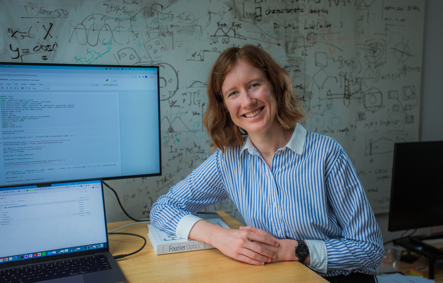 A smiling person with short, light brown hair sits at a desk with a computer monitor and laptop displaying code. Behind them is a whiteboard covered in mathematical formulas and diagrams. A book titled “Fourier Optics” is on the desk.