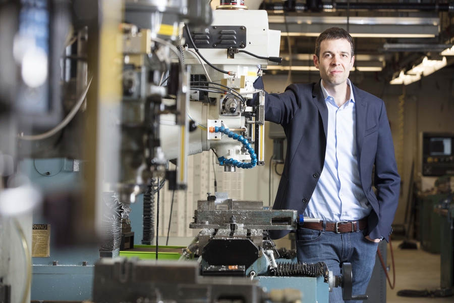 A man in a suit stands confidently next to industrial machinery in a workshop, with equipment and tools visible in the background. The setting appears modern and well-lit.