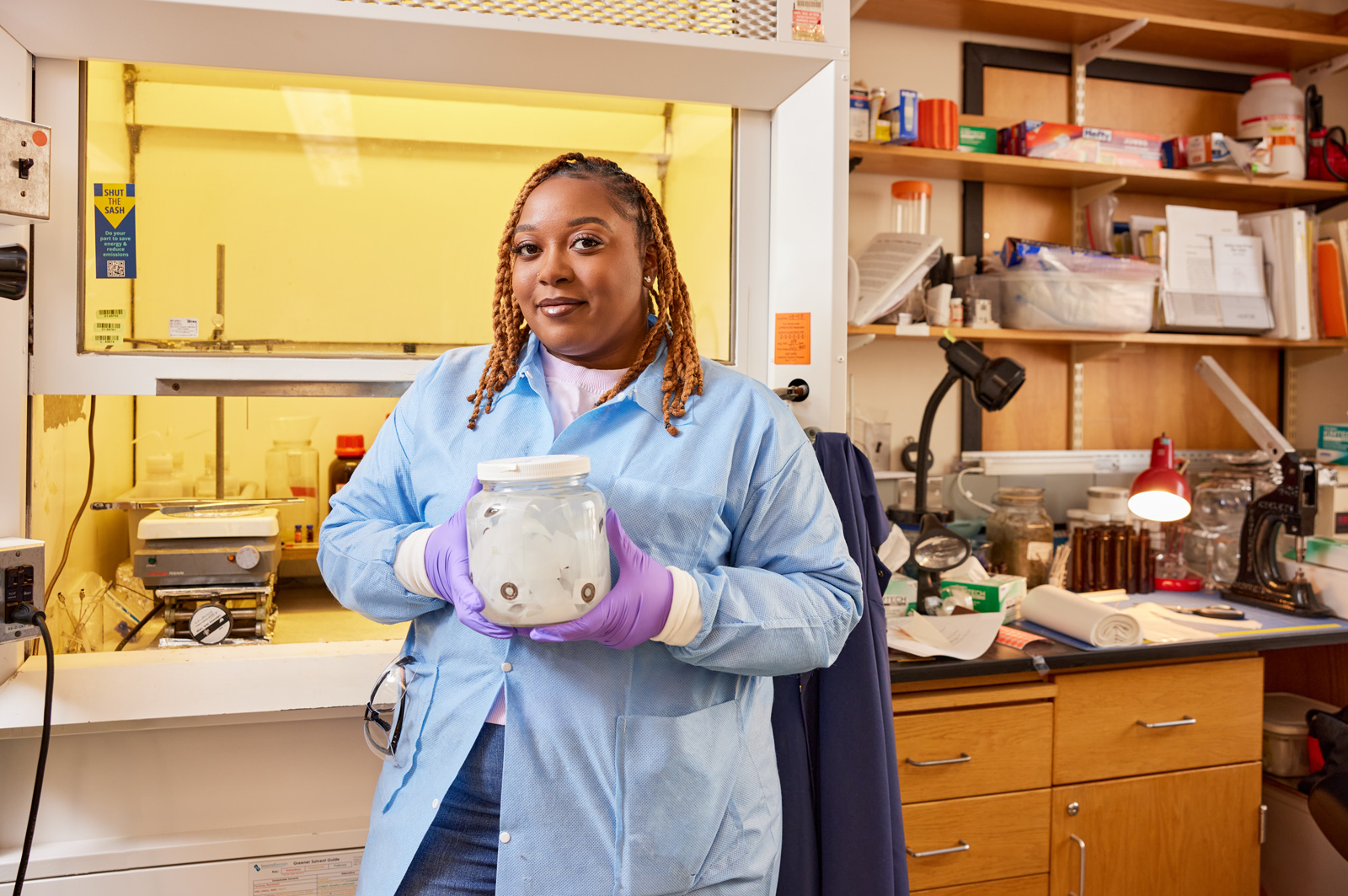 A woman in a lab coat and gloves stands in a laboratory holding a clear jar with small objects inside, in front of lab equipment and shelves with supplies.
