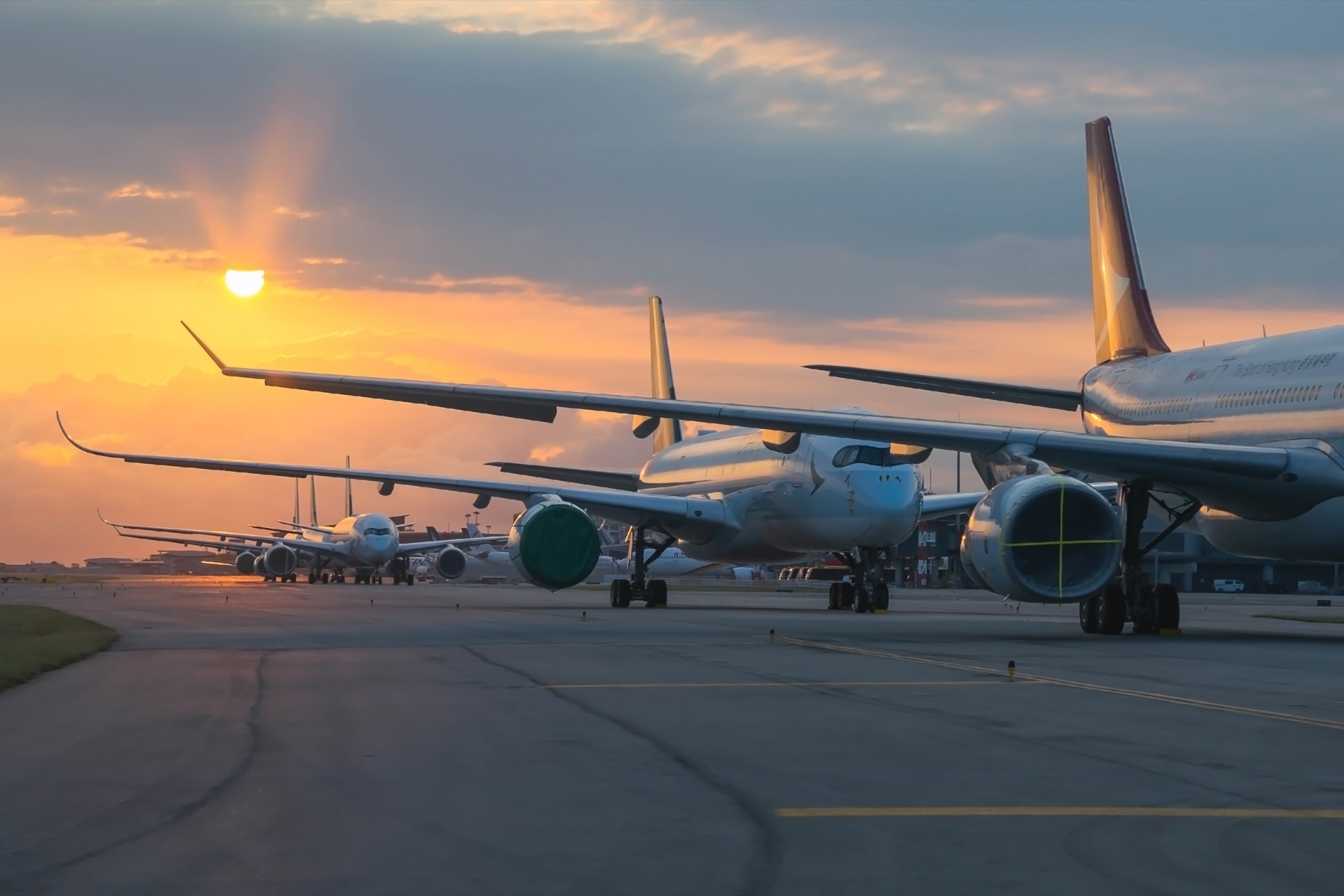 Several airplanes are parked in a row on an airport tarmac at sunset, with the sky glowing orange and yellow and clouds partially obscuring the sun. The planes’ tails and wings are silhouetted against the colorful sky.