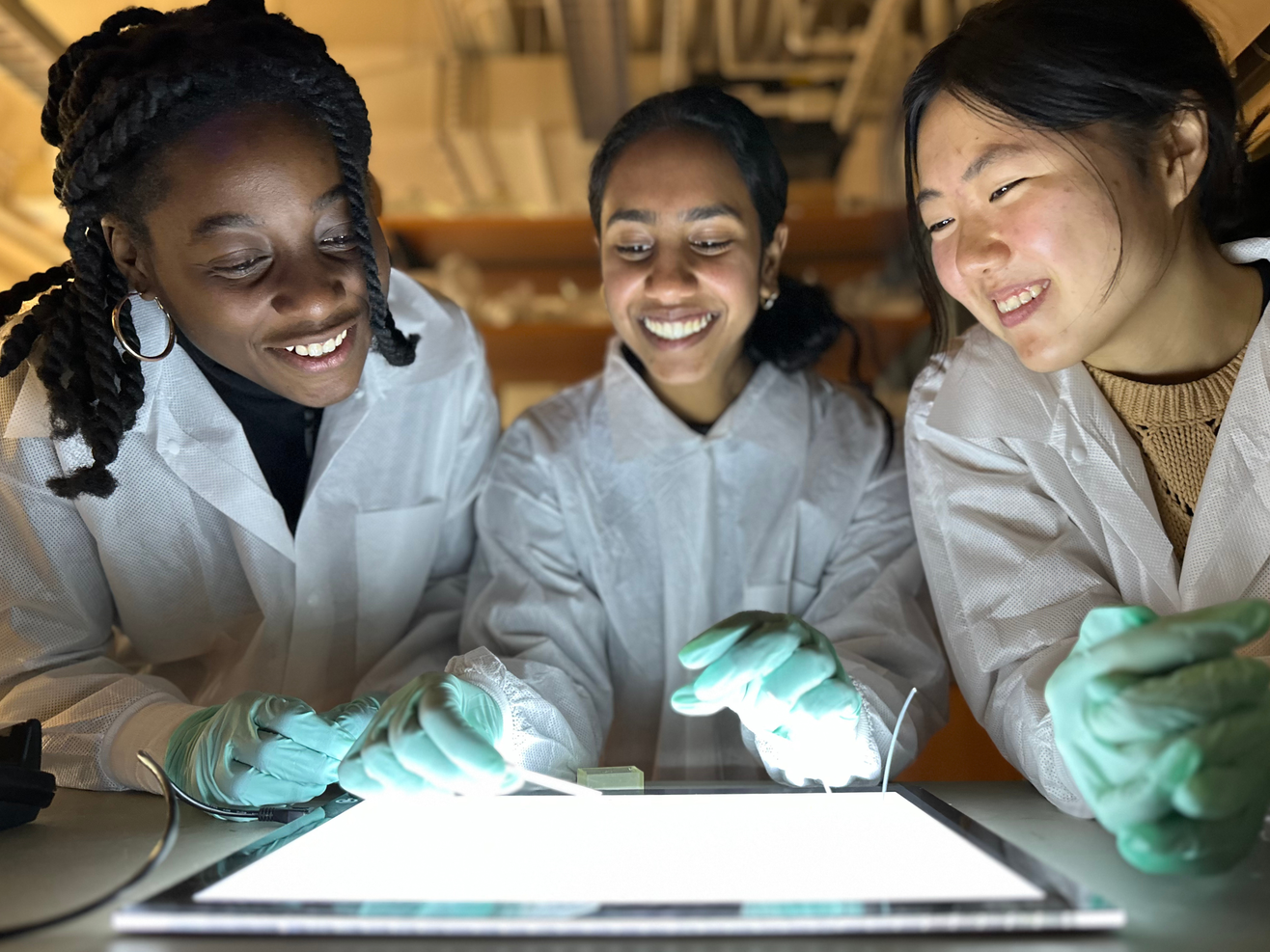 Three young women in white lab coats and turquoise latex gloves smile while conducting a laboratory experiment together. They are gathered around a white work surface with microscope slides, working collaboratively in a well-lit scientific workspace with blurred laboratory equipment visible in the background.