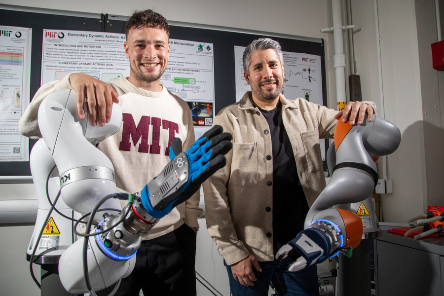 Two smiling people stand between two large robotic arms in a lab, with research posters on the wall behind them. One person wears an MIT sweater; the other wears a beige jacket.