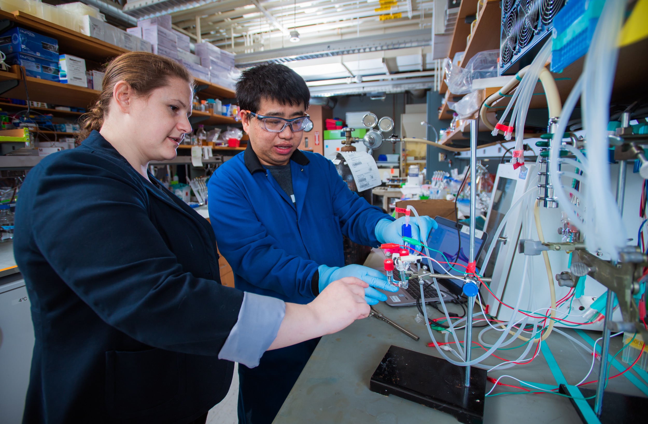 Two scientists in a lab coat and gloves work together on a complex experiment involving tubes, wires, and lab equipment, surrounded by shelves with various scientific supplies.