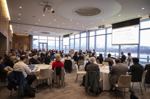 A large group of people sit at round tables in a modern conference room with floor-to-ceiling windows, listening to a panel discussion at the front. A large screen displays a presentation.