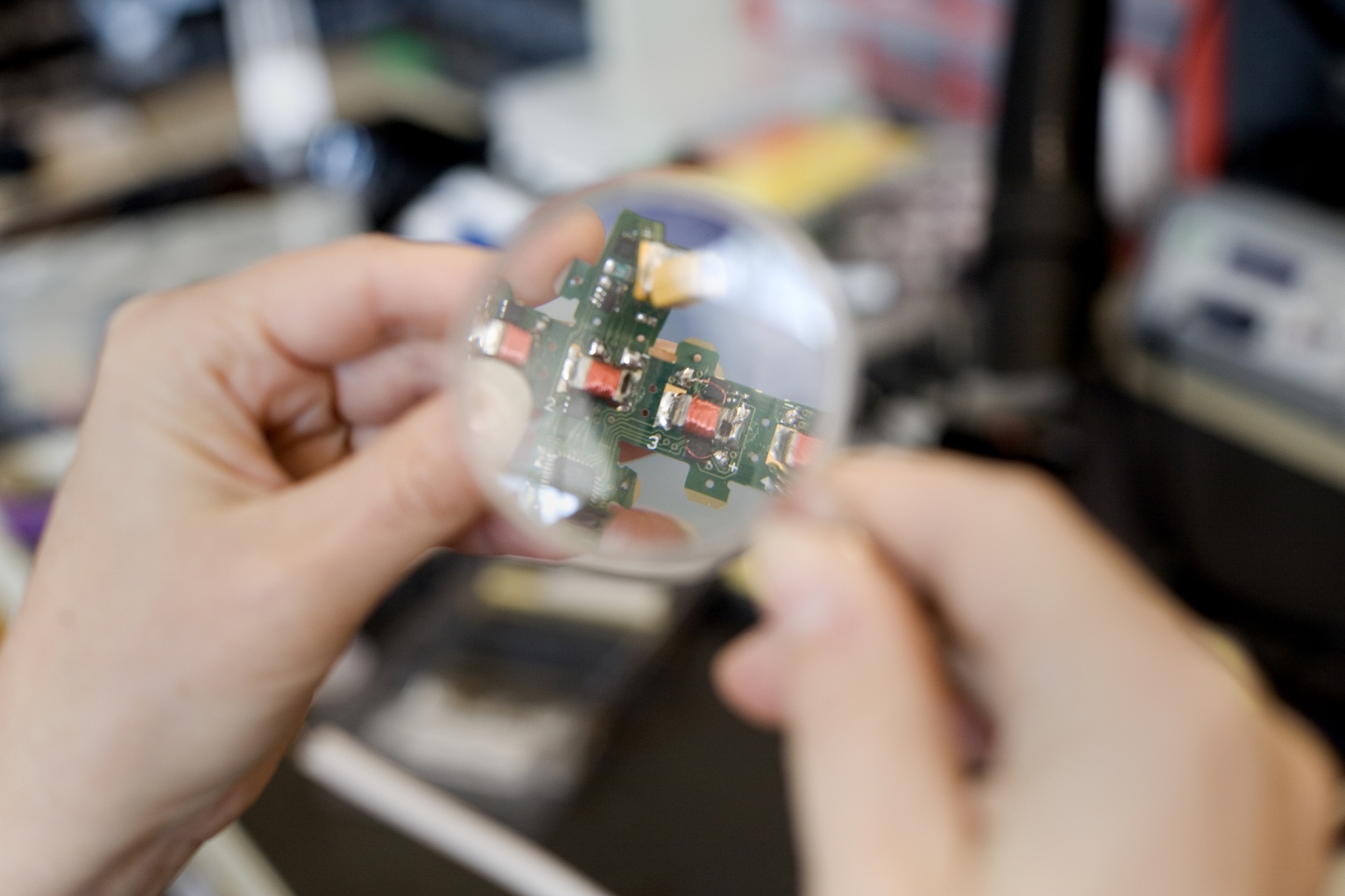 A person holds a magnifying glass over an electronic circuit board, closely examining its components in a brightly lit workspace.