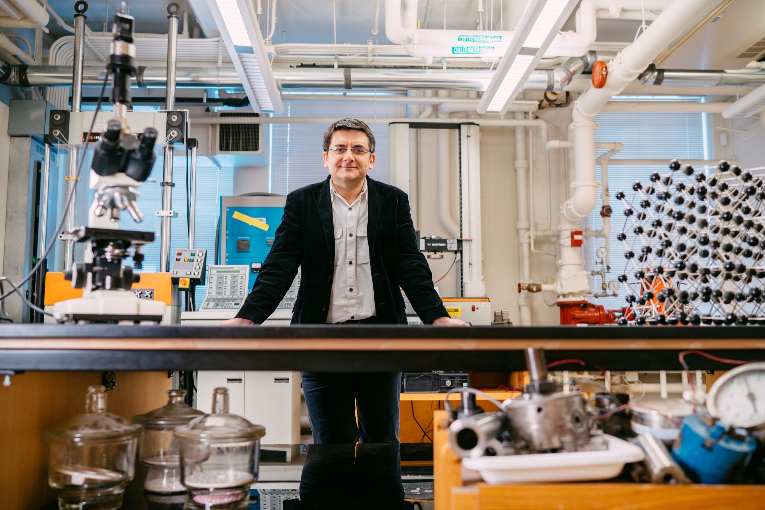 A man stands confidently behind a lab bench in a bright, modern laboratory filled with scientific equipment, glass containers, and machinery, with pipes and devices visible in the background.