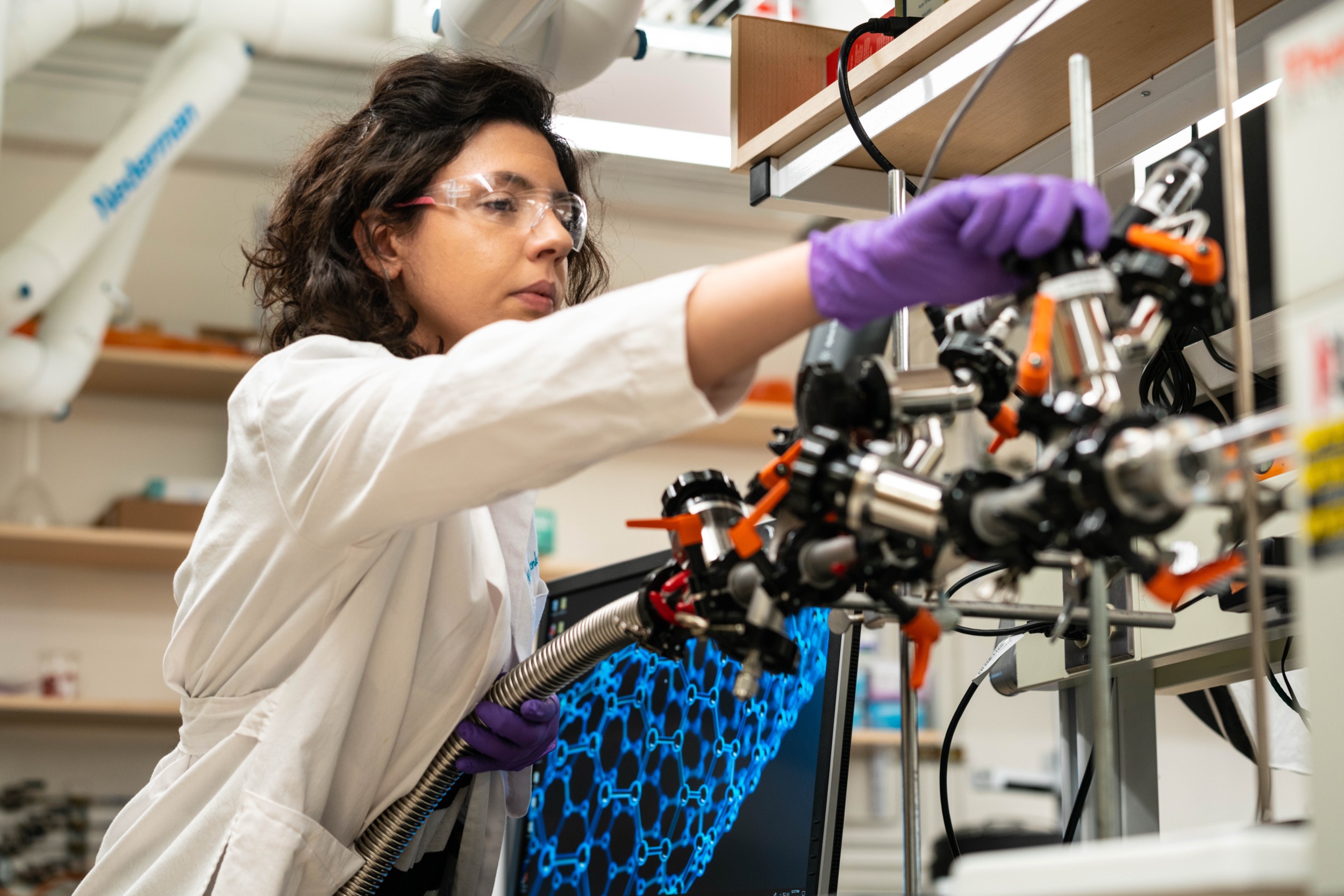 A scientist wearing safety goggles and purple gloves adjusts equipment in a laboratory, with a computer screen displaying a molecular structure in the background.