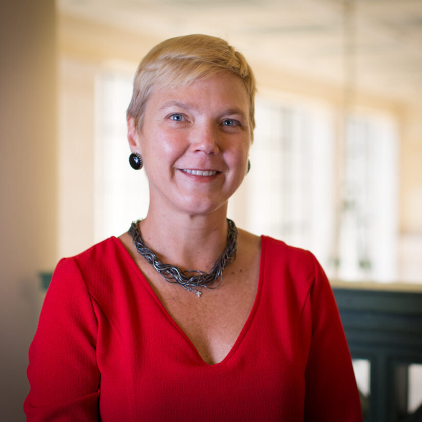 A woman with short blonde hair, wearing a red V-neck top, a chunky necklace, and black earrings, smiles at the camera in a bright, softly focused indoor setting.