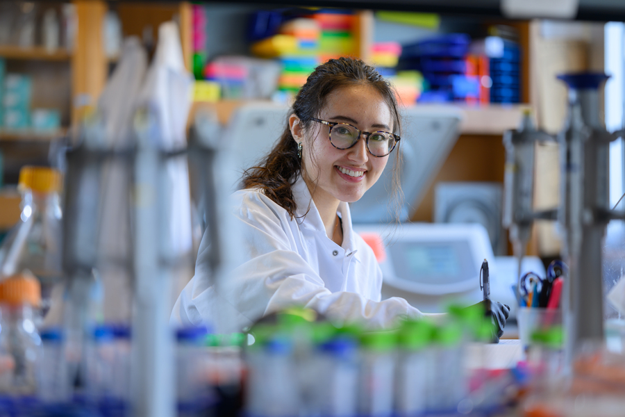 A woman wearing glasses and a white lab coat smiles while working in a laboratory, surrounded by scientific equipment and colorful lab supplies.