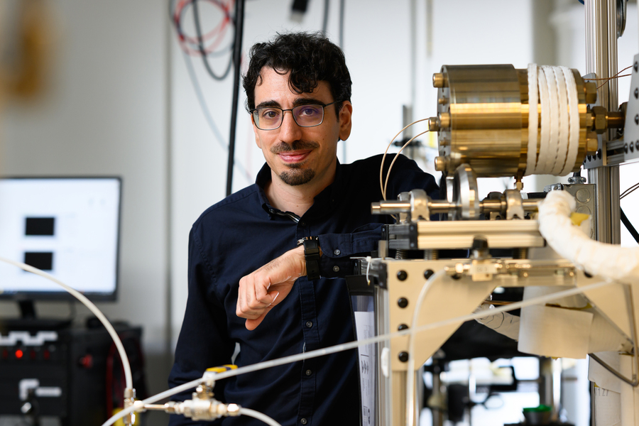 A man with glasses and dark curly hair smiles while standing in a laboratory, leaning on scientific equipment with wires and metal components. A computer monitor and lab instruments are visible in the background.