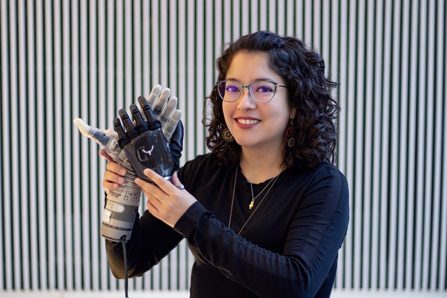 A woman with curly hair and glasses smiles while holding and adjusting a robotic prosthetic hand. She stands in front of a background of vertical black and white stripes.