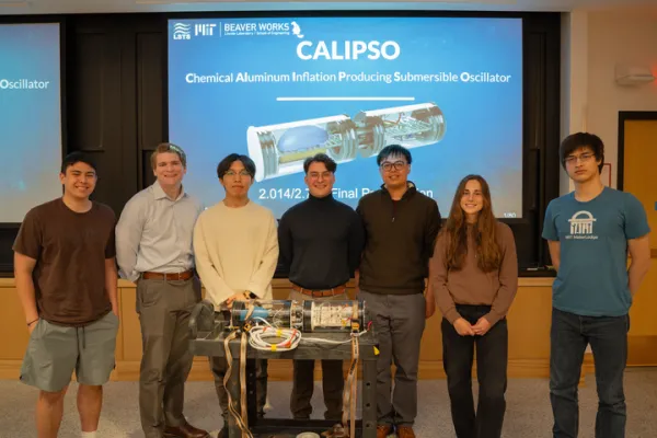 Seven people stand in front of a presentation screen displaying “CALIPSO Chemical Aluminum Inflation Producing Submersible Oscillator.” They pose with a technical device on a table in a classroom setting.
