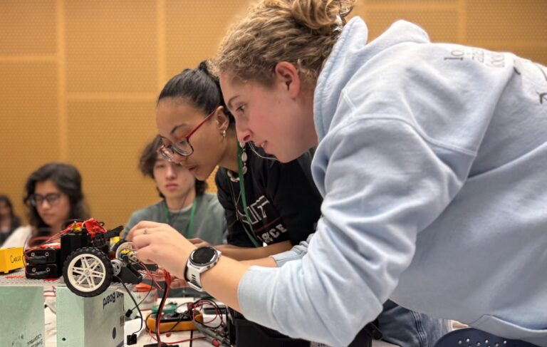 Two students focus intently as they work together on assembling or repairing a small robotic vehicle, surrounded by electronic components, with other students visible in the background.