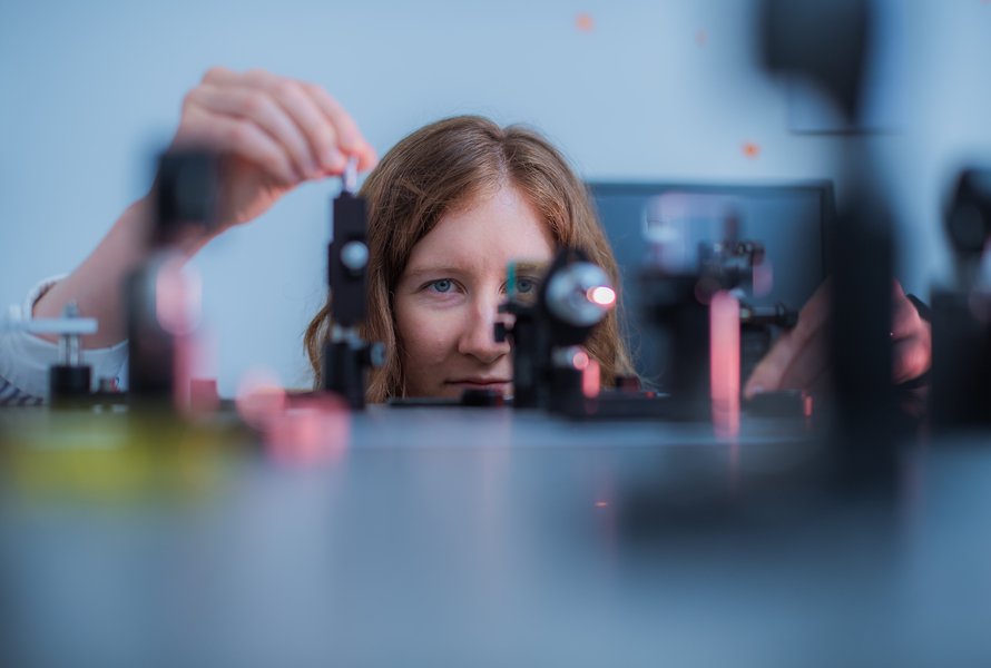 A person with long brown hair adjusts scientific equipment on a laboratory table, focusing intently on the setup, with various optical instruments and lenses visible in the foreground.