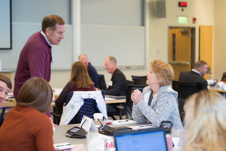 A man in a maroon sweater leans over to speak with a seated woman in a gray jacket in a bright classroom, while other people work and converse in groups at tables in the background.