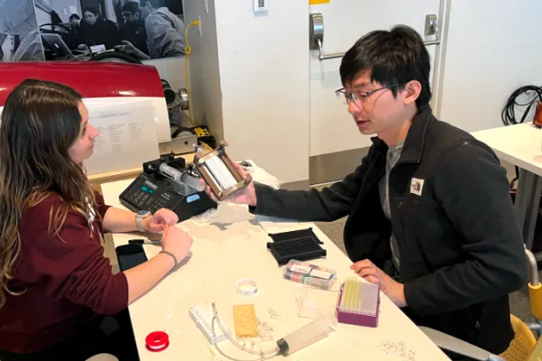 Two people sit at a table in a lab, examining scientific equipment. One holds a device while the other observes. Various lab tools, pipette racks, and supplies are on the table. A black-and-white photo hangs on the wall behind them.