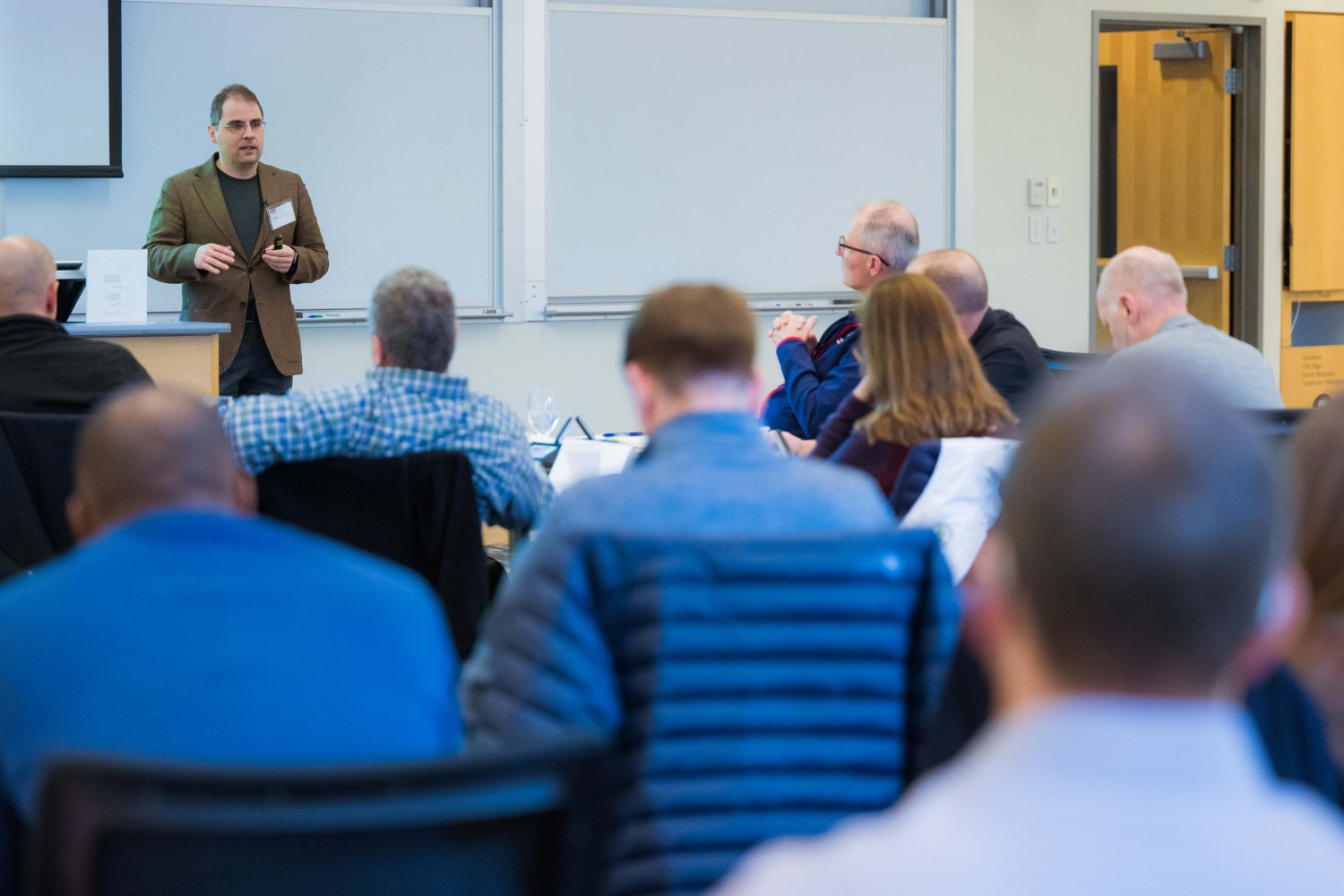 A man stands at the front of a classroom giving a presentation to an audience of seated adults, who are facing him and listening attentively. Whiteboards and a door are visible in the background.