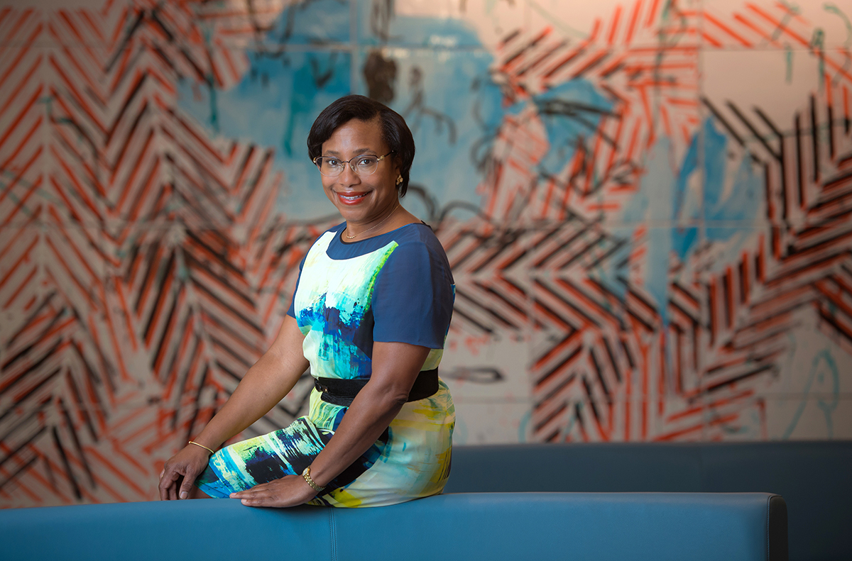 A woman with short dark hair, glasses, and a colorful dress sits on a blue couch, smiling at the camera. The background features an abstract wall mural with blue, black, and orange patterns.