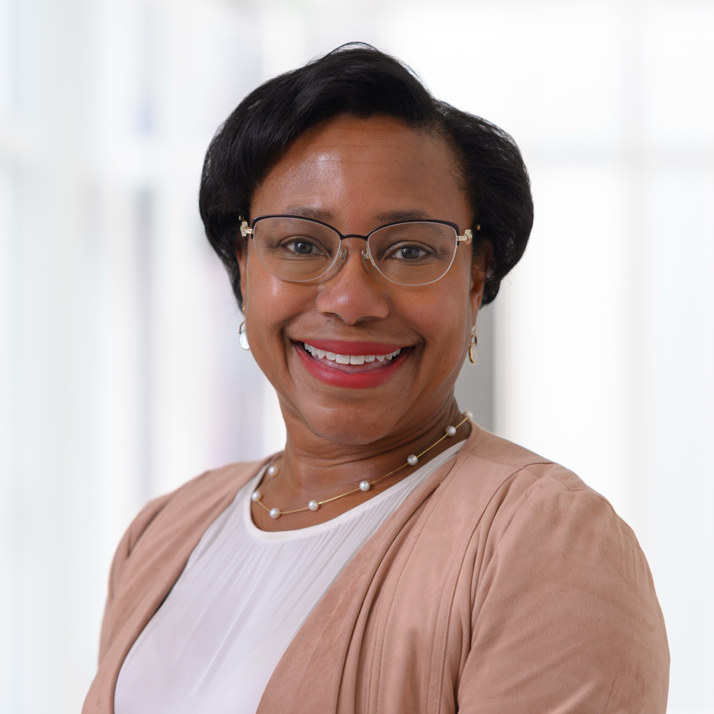 A woman with short black hair, wearing glasses, a white top, a light brown blazer, and a pearl necklace, smiles at the camera in a bright, blurred indoor setting.