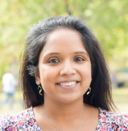 A woman with long dark hair smiles at the camera outdoors. She is wearing earrings, a necklace, and a patterned top. The background is blurred greenery.