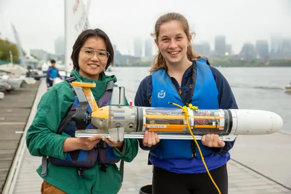 Two young women wearing life jackets stand on a dock by the water, smiling and holding a small underwater robot or submersible. Boats and a city skyline are visible in the background under a cloudy sky.