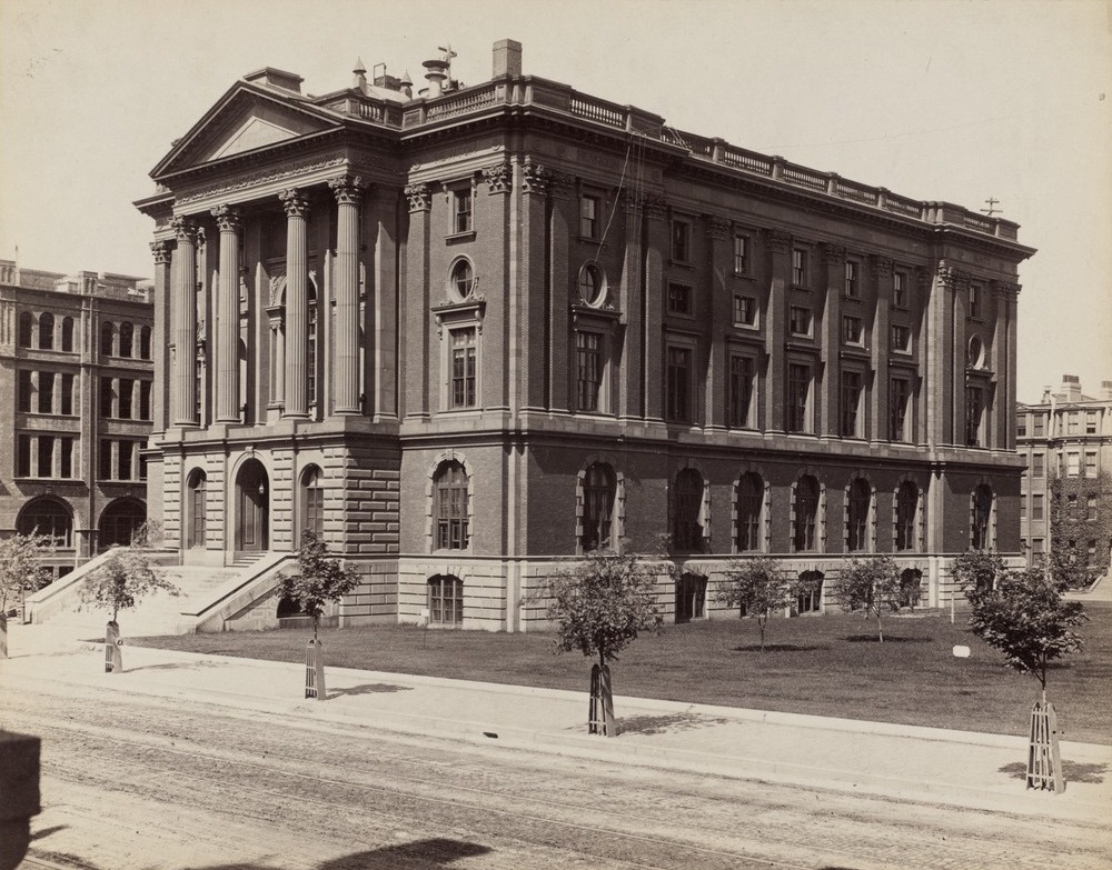 Historic black-and-white photo of a large, classical-style brick building with tall columns, arched windows, and wide steps leading to the entrance, surrounded by a lawn and young trees.