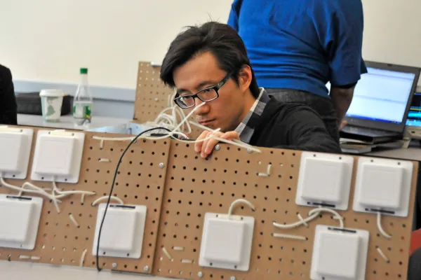 A person wearing glasses works intently with wires on a pegboard setup with several white devices attached. A laptop and another person are visible in the background.