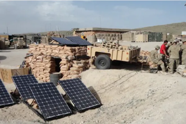 A military outpost with sandbag fortifications and several solar panels set up on the ground. Soldiers stand nearby, and various equipment and trailers are visible in the background.