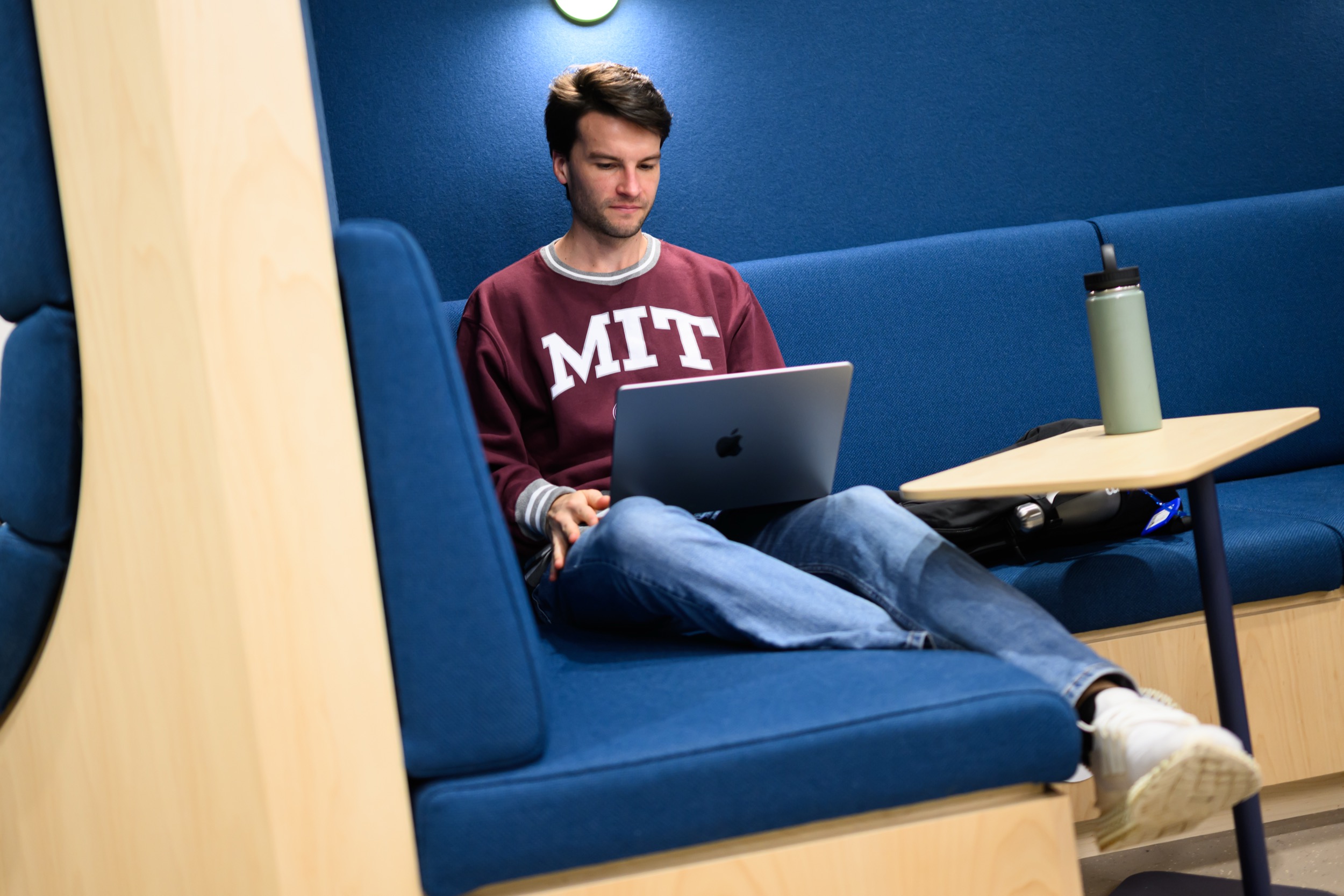 A person in an MIT sweatshirt sits on a blue cushioned bench with a laptop on their lap, looking at the screen. A green water bottle and a backpack are on the nearby table.